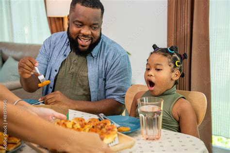 African family parents and two little daughter eating fried chicken and pizza on table for ...