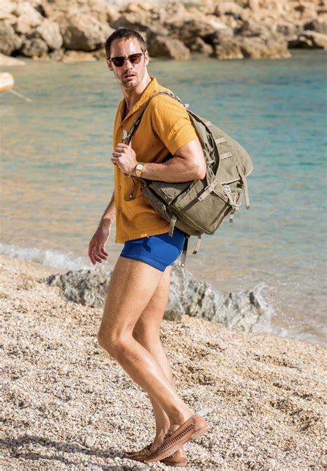 a man walking on the beach with a backpack in his hand and water behind him