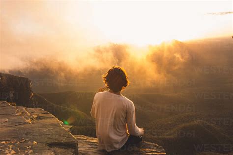 Rear View Of Man Sitting On Cliff While Looking At Landscape During ...