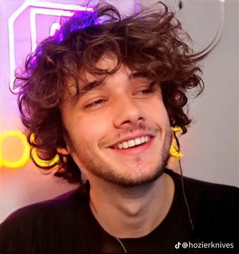 a young man with curly hair smiles into the camera while wearing headphones in front of a neon sign