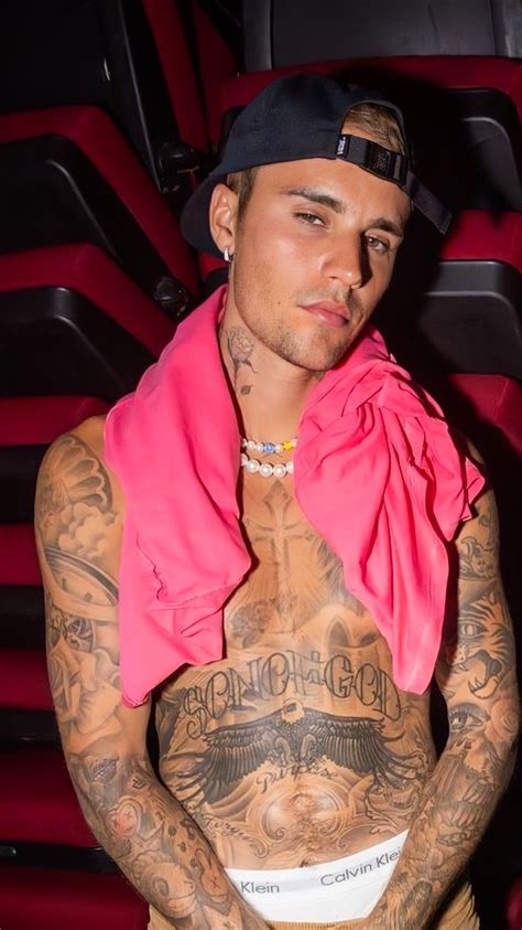 a man with tattoos on his chest wearing a pink shirt and black hat, standing in front of red chairs