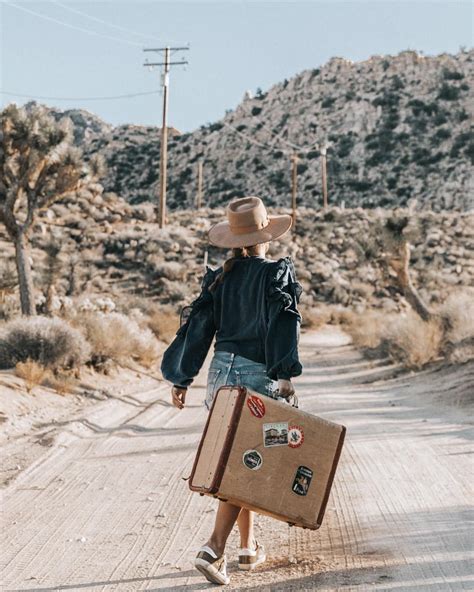 a woman walking down a dirt road with a suitcase in her hand and a hat on top of her head