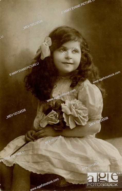 A little girl in a frilly white dress, holding a bunch of artificial roses in the photographer's ...