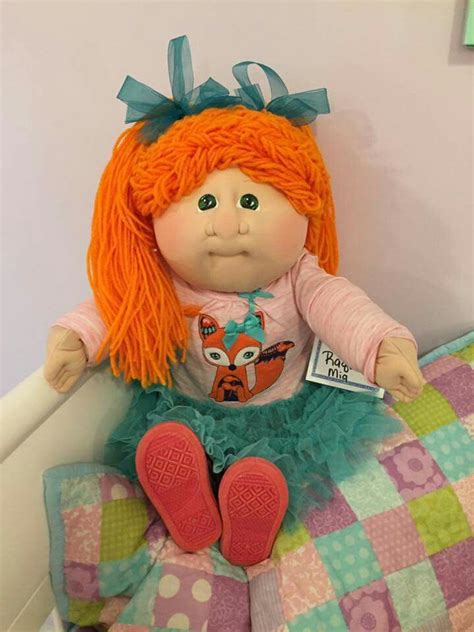 an orange haired doll sitting on top of a bed next to a pink and green pillow