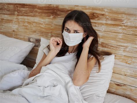 woman in medical mask lying in bed under the covers and gesturing with