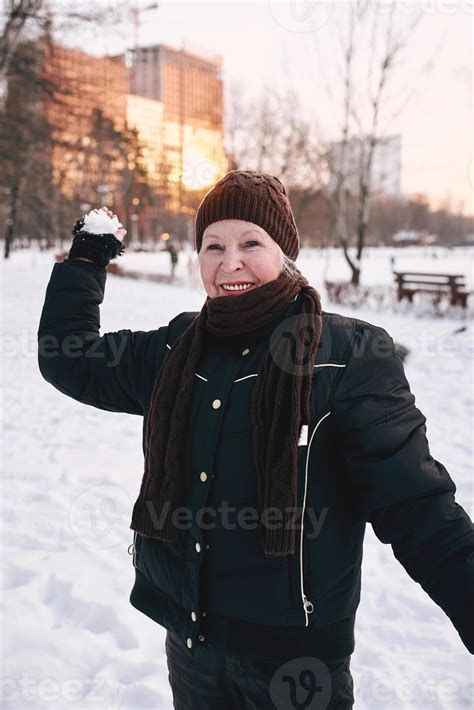 senior woman in hat and sporty jacket snowballing in snow winter park