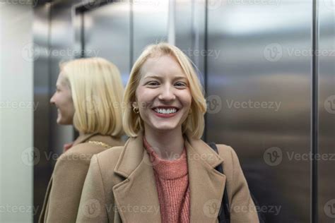Cute blond female in trendy coat staying in lift alone and smiling