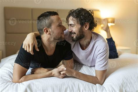 Happy gay couple lying down on the bed at home, hugging and flirting