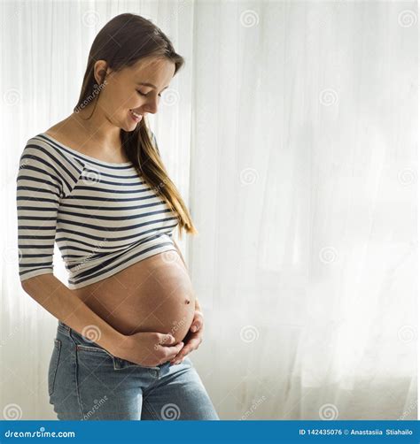 Happy Attractive Pregnant Woman Standing Near the Window and Holding