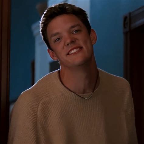 a smiling young man wearing a tan sweater in front of a blue wall and doorway